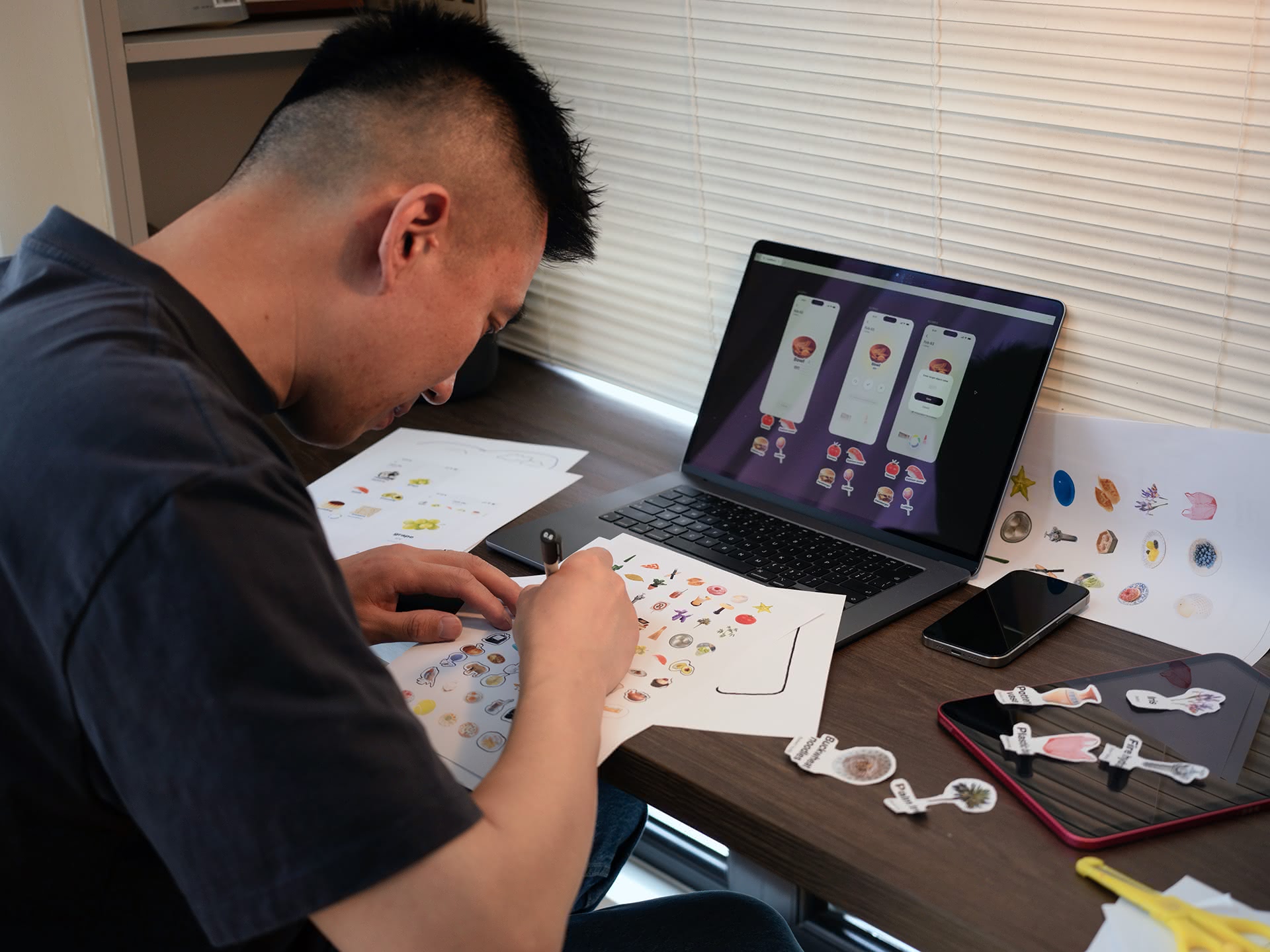A photo of a man sketching stickers on a piece of white paper at a desk. Before him is a MacBook showing images from the app CapWords.
